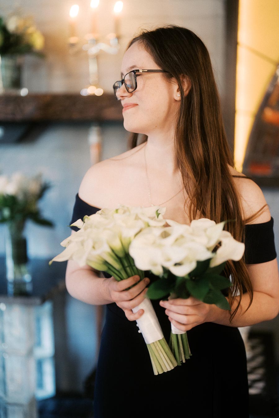 Young woman in a black off-shoulder dress holding a bouquet of white flowers, smiling and standing indoors with blurred candlelit background. Perfect for wedding or celebration themes.