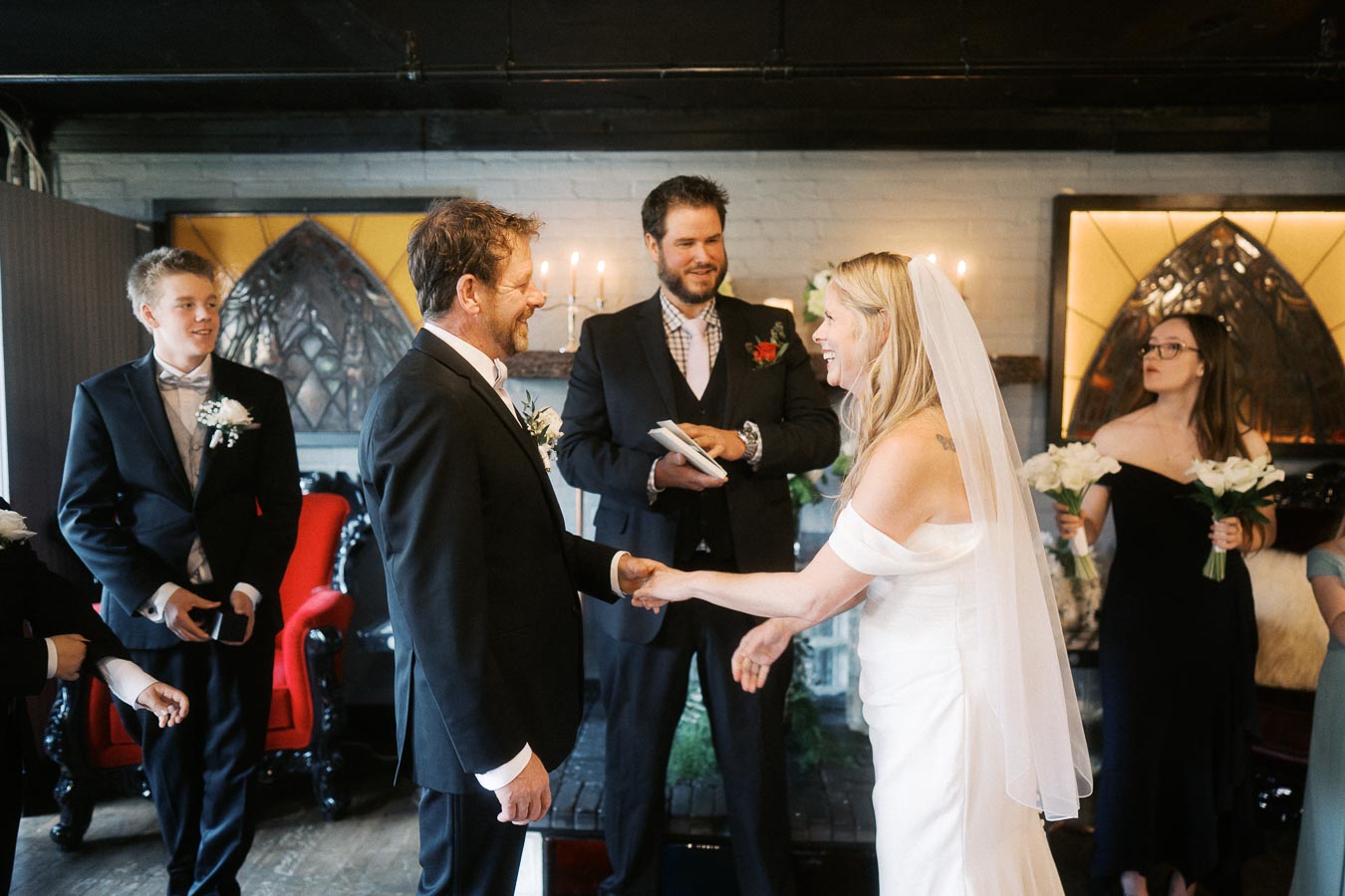 A bride and groom exchanging vows in an intimate wedding ceremony, surrounded by their wedding party, with an officiant present in a warmly lit venue.