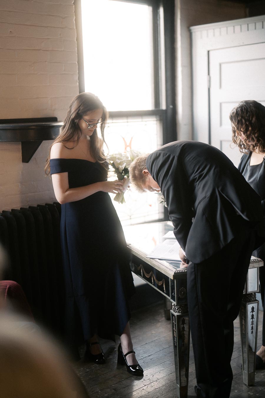 Three people engaged in a wedding ceremony, with a woman holding a bouquet and a man signing a document in a dimly lit room.