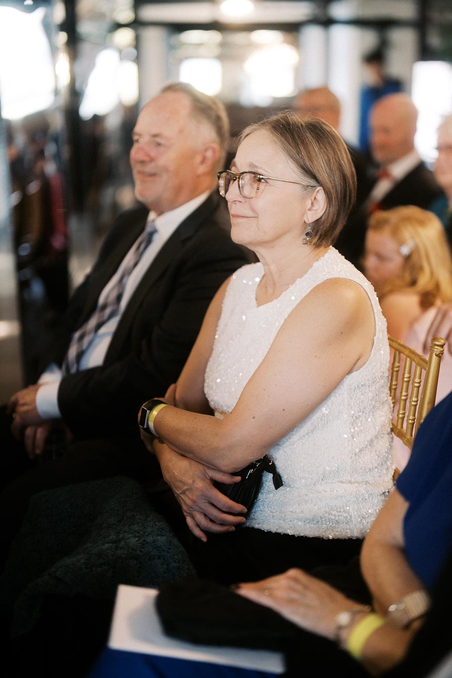 Elderly couple sitting together at an indoor event, with the woman wearing a white sequined top and glasses, both appearing attentive and thoughtful.