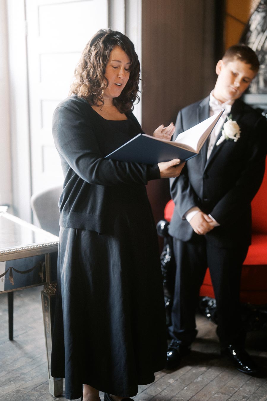 Woman in formal attire reading from a large book, standing next to a young boy in a suit with a white flower corsage, in an elegantly decorated room.