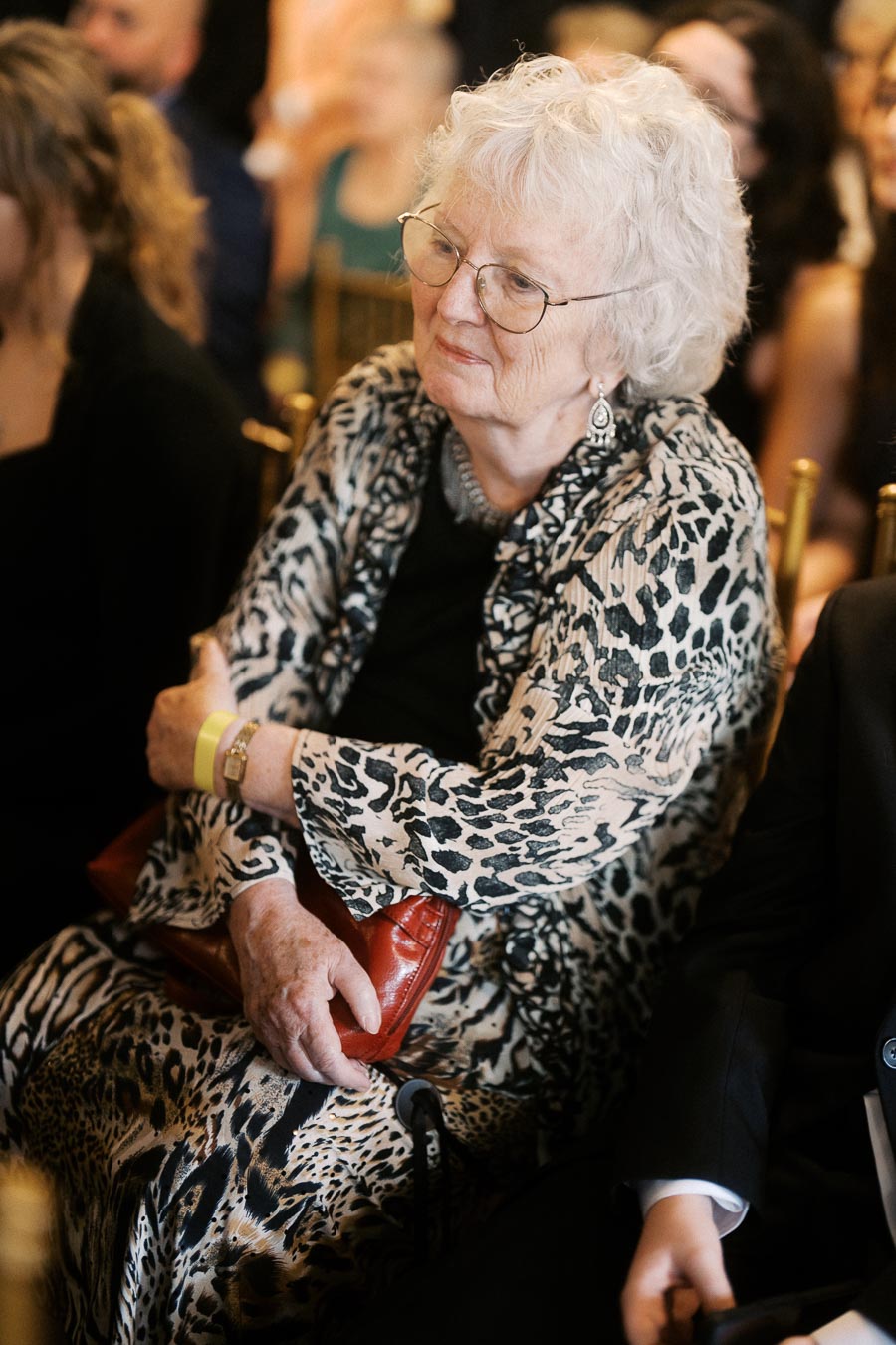 Elderly woman with white hair wearing a leopard print outfit, seated and holding a red clutch, attending an event.