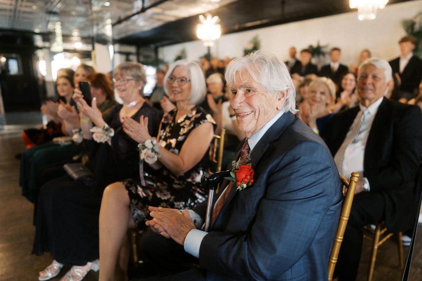 Elderly man in a suit smiling and applauding at a formal indoor event, surrounded by a group of people in elegant attire, creating a joyful atmosphere.