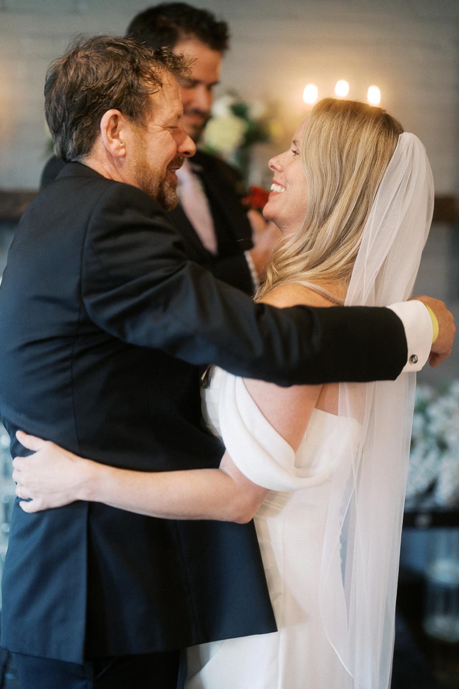 A bride and groom embracing joyfully during their wedding ceremony, with the bride in a white gown and veil, set in an elegantly decorated venue with soft lighting and floral arrangements in the background.