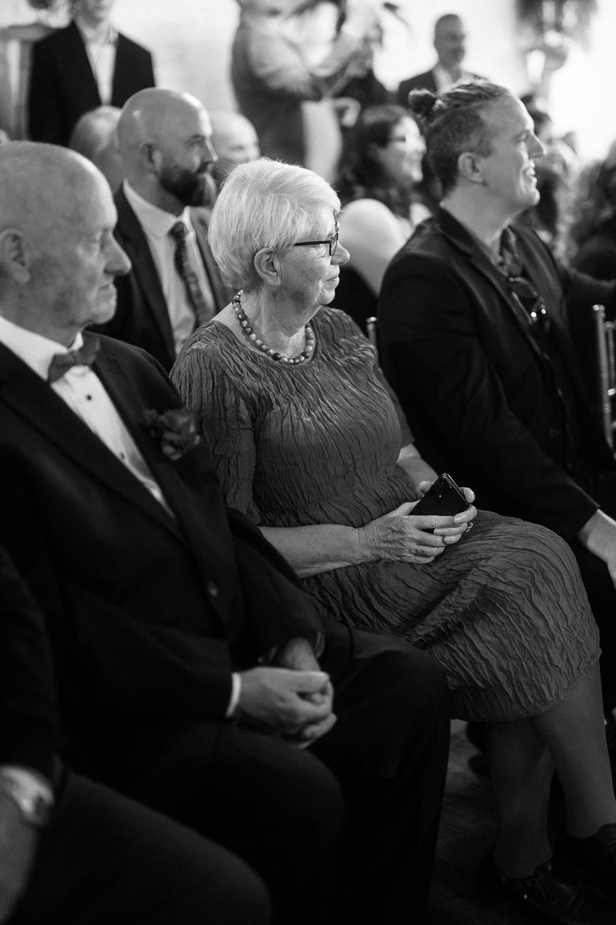 Black and white photo of attendees sitting at a formal event, including an older woman with glasses and a smartphone, and men in suits.