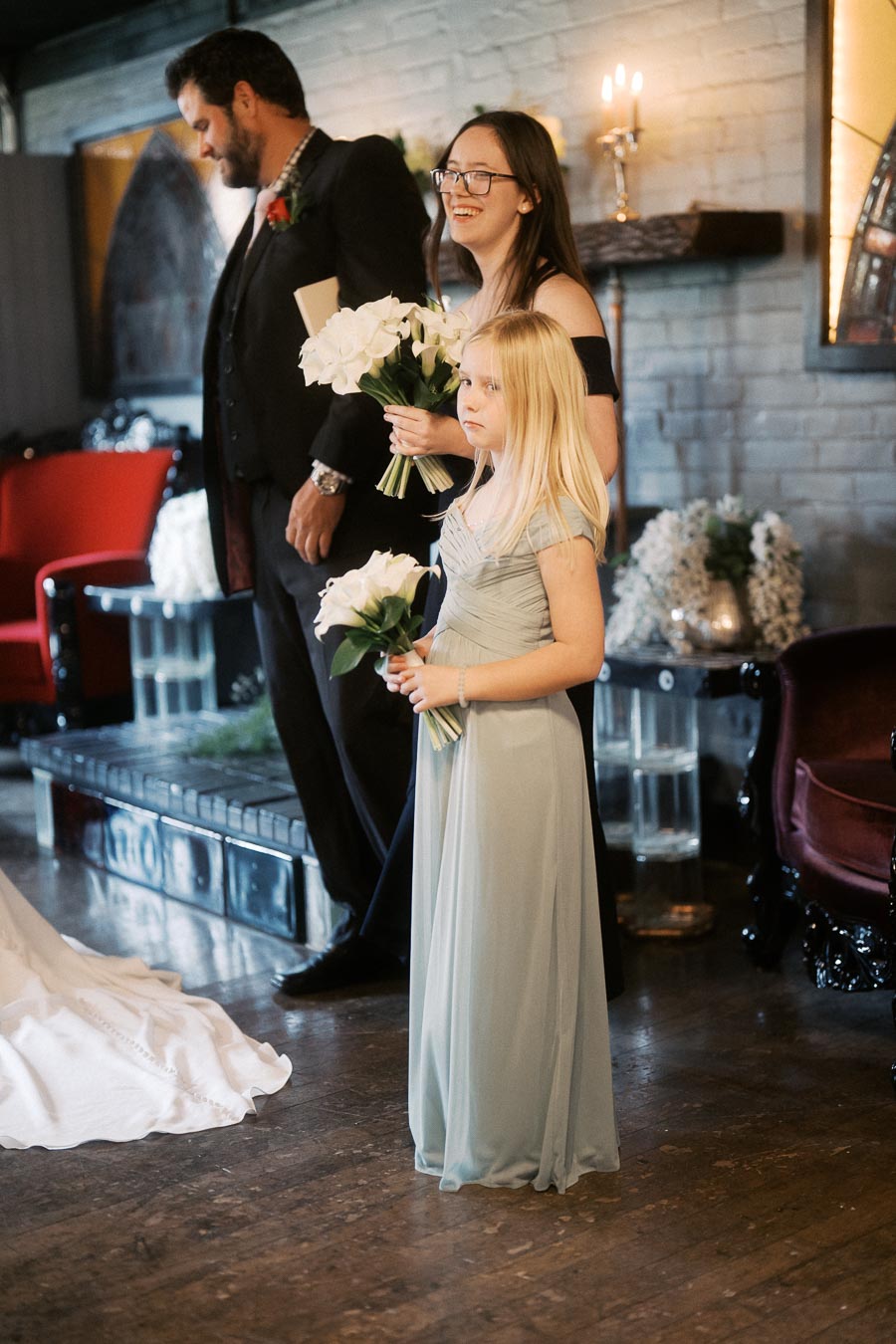 Bridesmaids and groomsman in elegant attire holding bouquets at an indoor wedding ceremony.