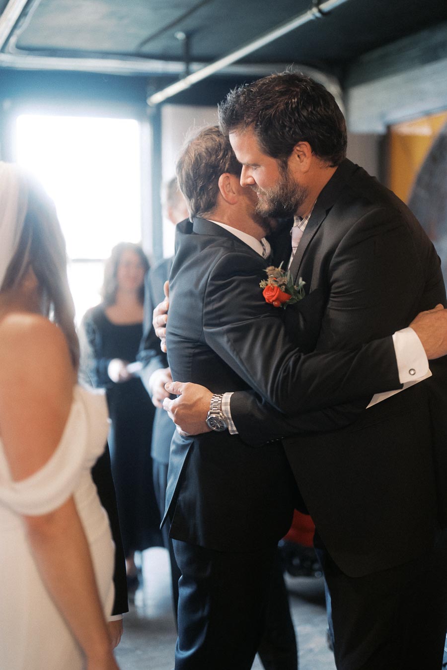 Groom and best man sharing an emotional hug at a wedding ceremony, with the bride visible in the foreground, wearing an elegant white dress.