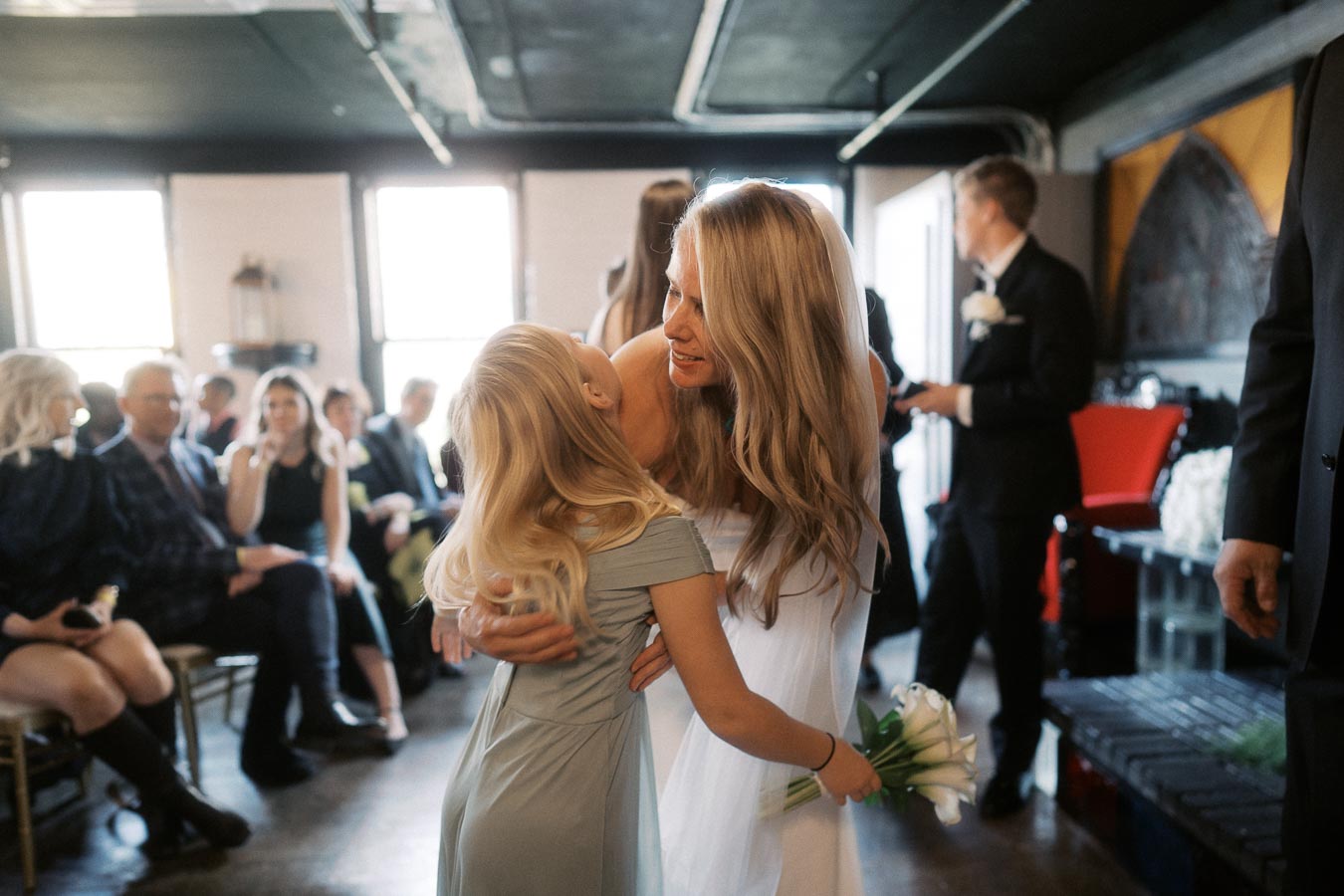 A bride smiling and embracing a young flower girl in a warmly lit wedding ceremony venue, with guests seated in the background.