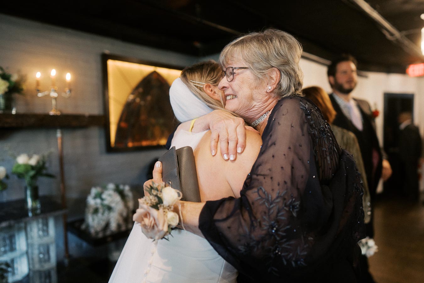 Elderly woman warmly embracing bride at indoor wedding reception, both smiling.