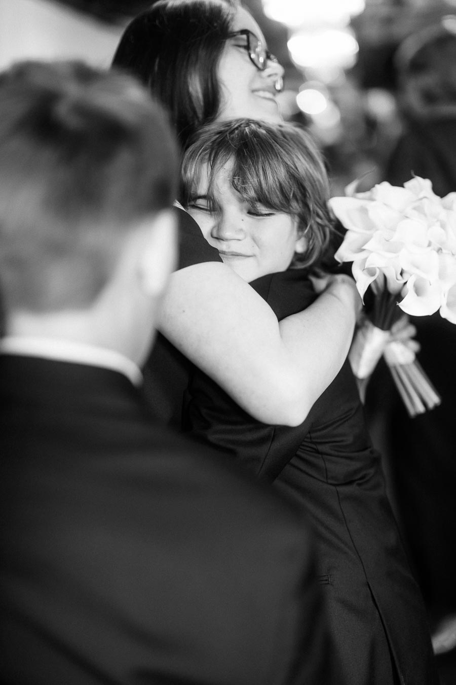 Black and white photo of a joyous embrace between a woman holding a bouquet and a young boy, both smiling, surrounded by elegantly dressed people at a formal event.