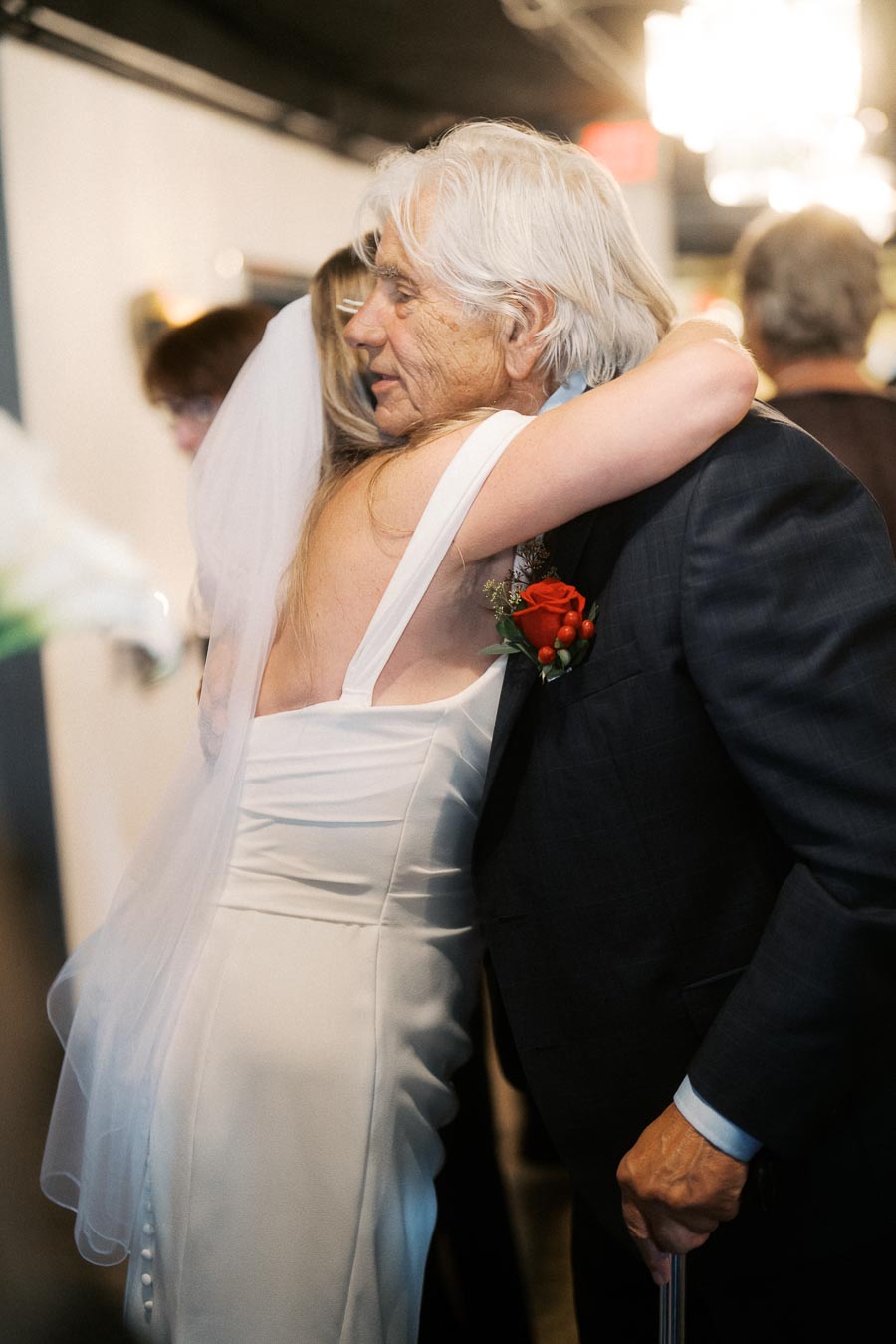 Elderly man in suit warmly hugging a bride in a white wedding dress with a veil indoors.