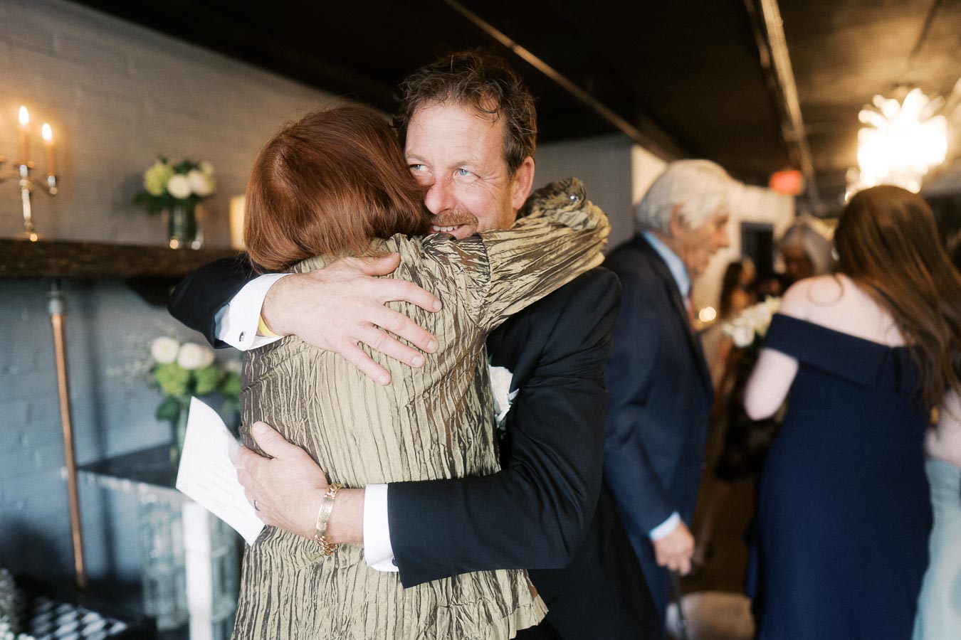 A man in a suit warmly hugging a woman in a gold jacket at a formal gathering, with blurred guests and elegant decor in the background.