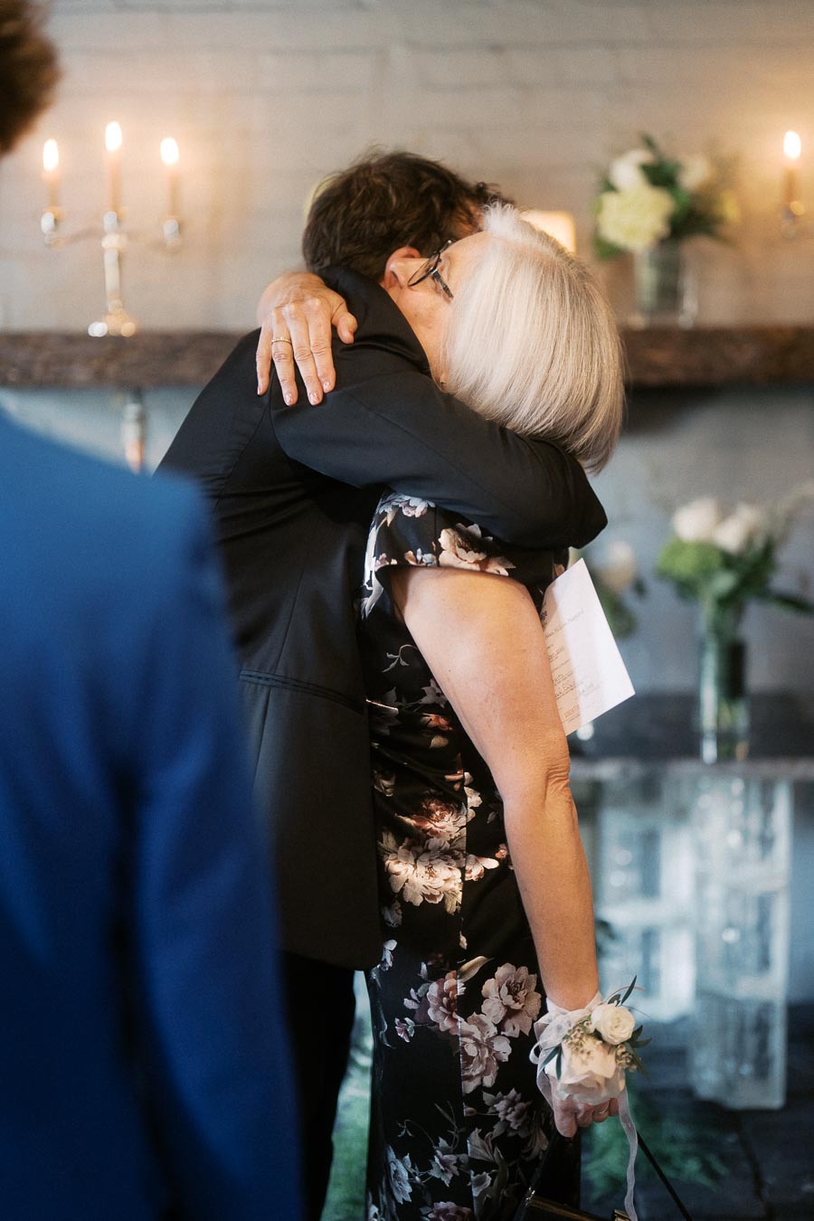An elderly woman in a floral dress with a corsage hugs a man in a dark suit during an intimate indoor ceremony illuminated by candlelight, evoking a sense of warmth and affection.