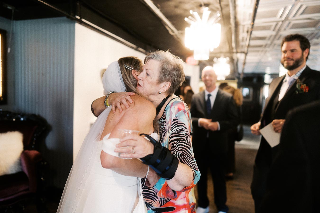 Elderly woman warmly embracing bride in wedding dress at indoor celebration, with guests smiling in the background.