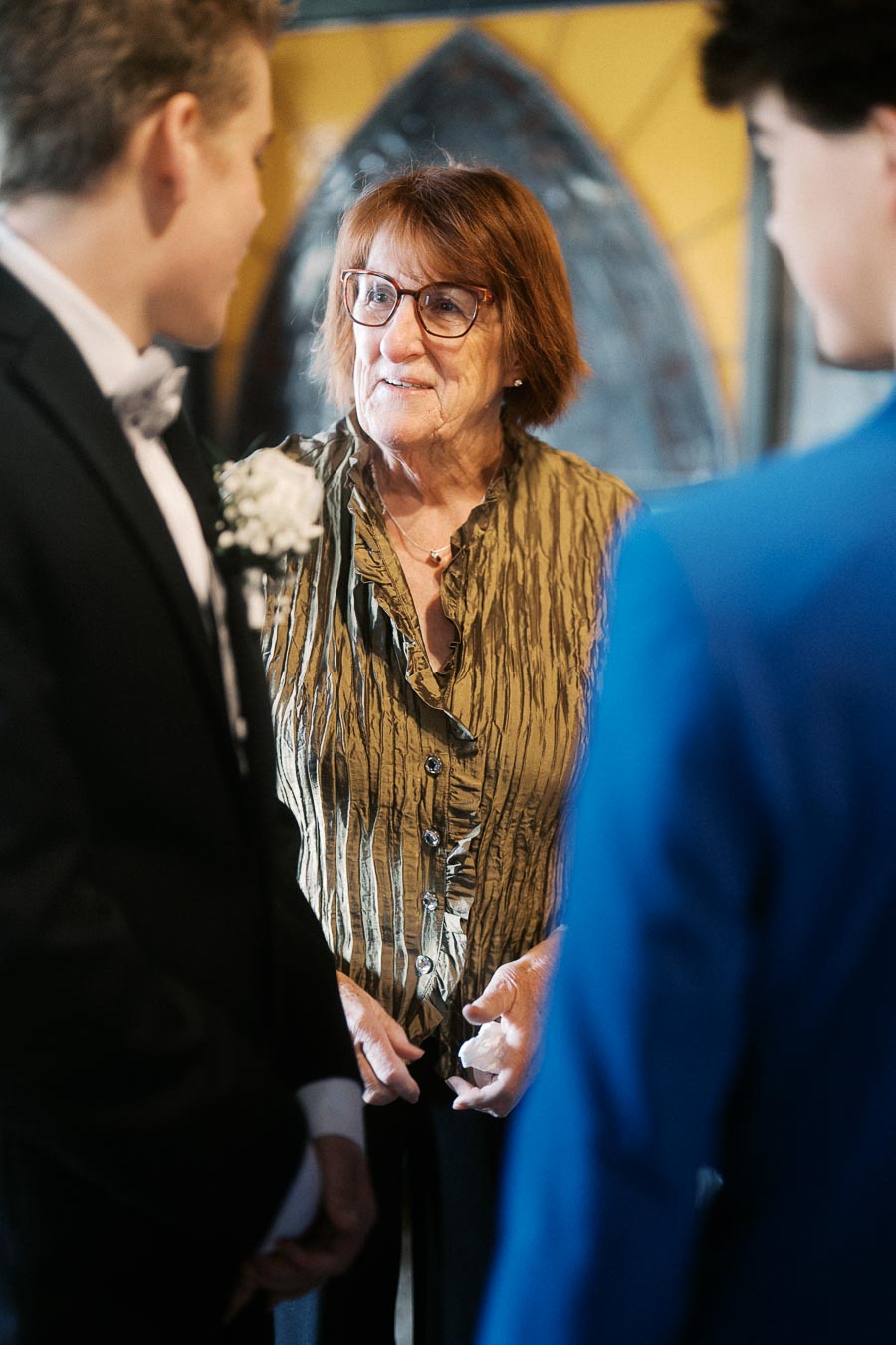 An elderly woman in glasses, wearing a green blouse, engaged in conversation with two young men dressed formally, one in a black suit and the other in blue, with a blurred stained glass window background.