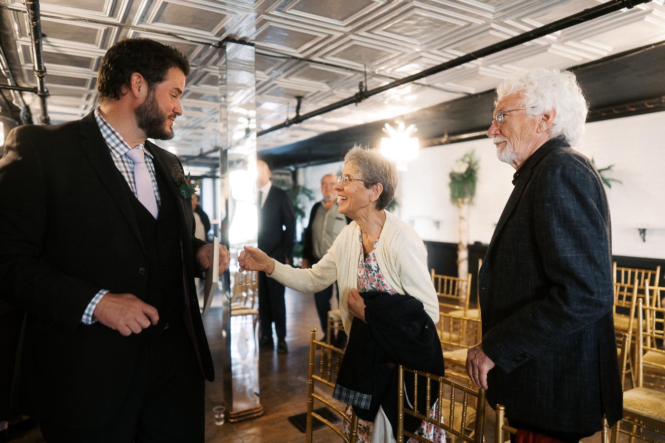 A man in a suit greets an elderly woman and man at an indoor event with golden chairs and elegant decor. The woman smiles warmly while extending her hand in greeting, highlighting a friendly interaction.