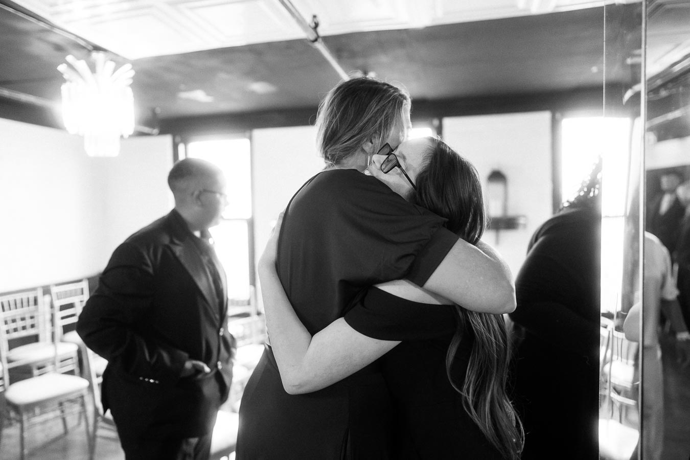 A black and white photo capturing an emotional hug between two people at a formal gathering, with another person in the background looking on. The setting appears to be indoors with elegant decor and seating arrangement.