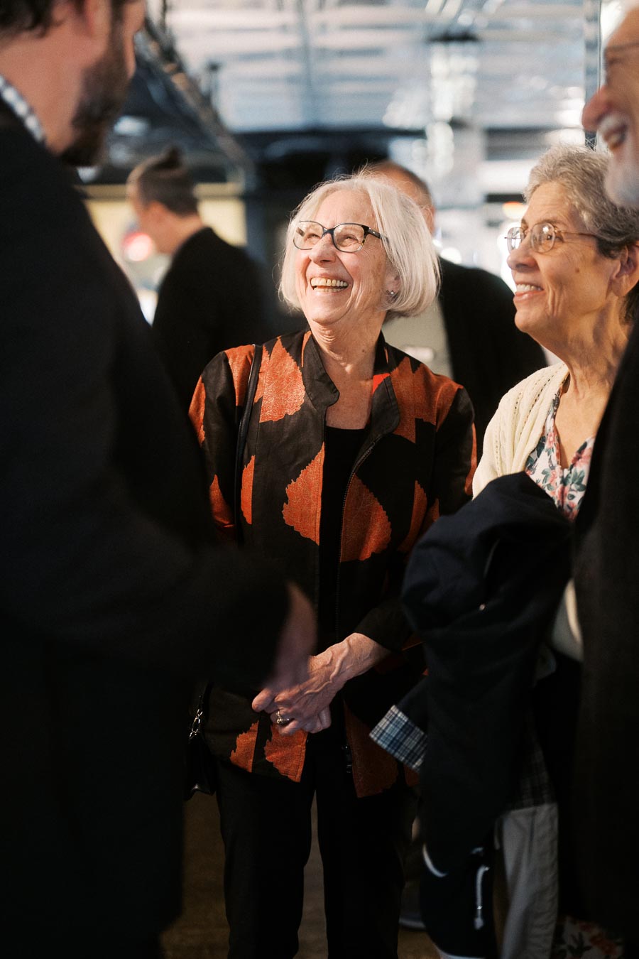 Elderly woman smiling and engaging in conversation at a social gathering, wearing glasses and a stylish jacket.