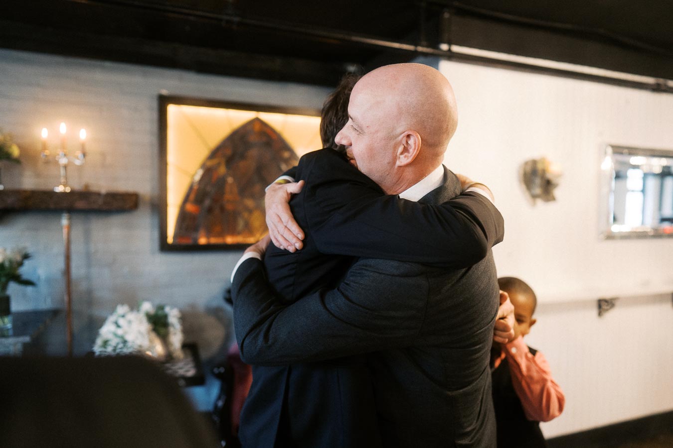 A bald man in a suit warmly embraces another person in a cozy, dimly lit room, with a child in the background. The setting includes a lit candle and decorative elements, suggesting a special occasion or event.