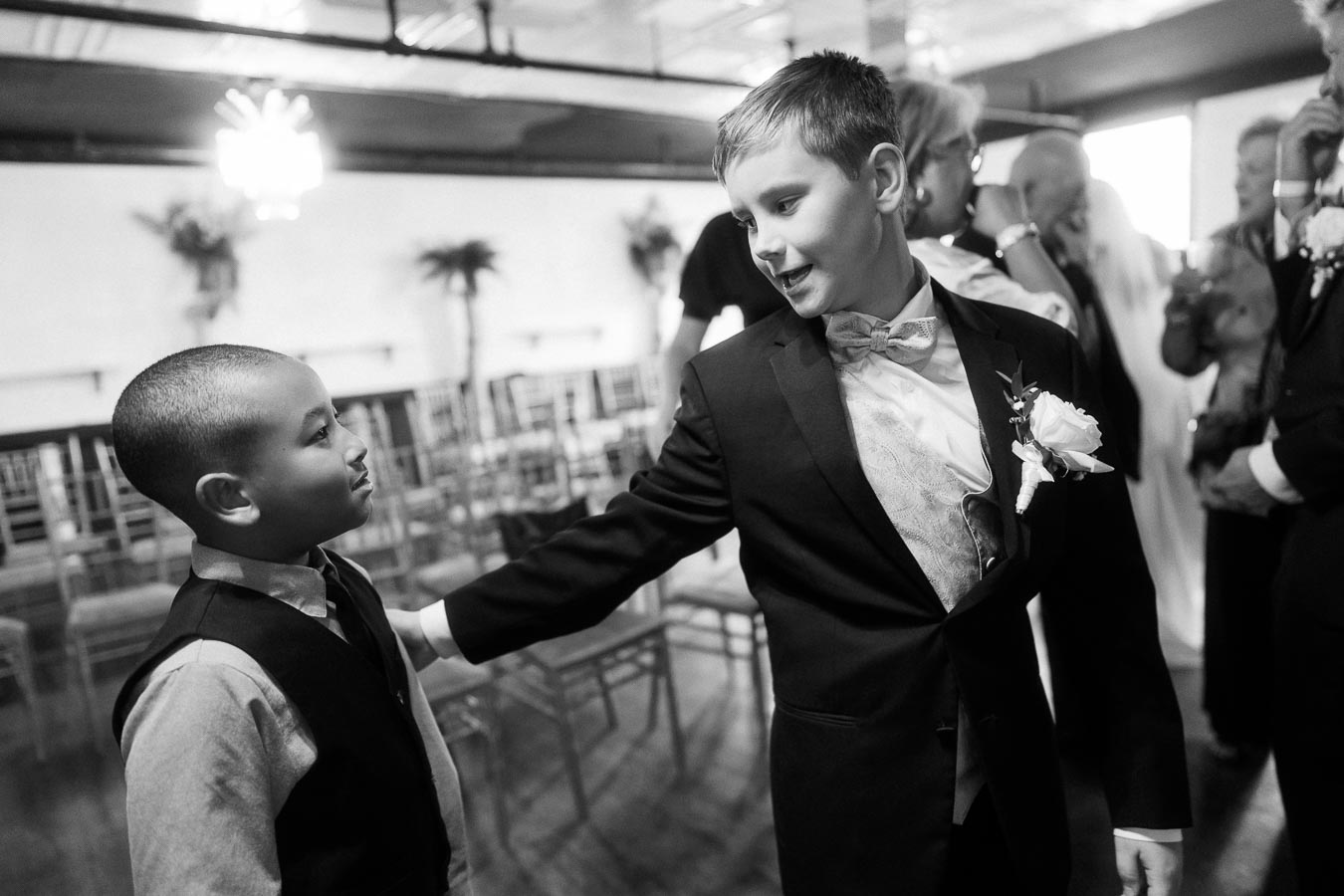 Black and white photo of two young boys dressed in formal attire at a wedding venue, smiling and interacting in a warm indoor setting with chairs and blurred guests in the background.