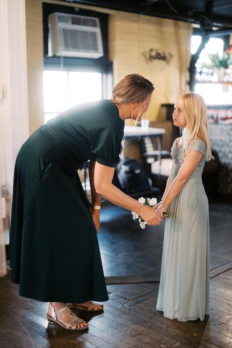 Woman in a green dress bending down to talk to a young girl in a light blue dress, while handing her a small flower bouquet indoors.