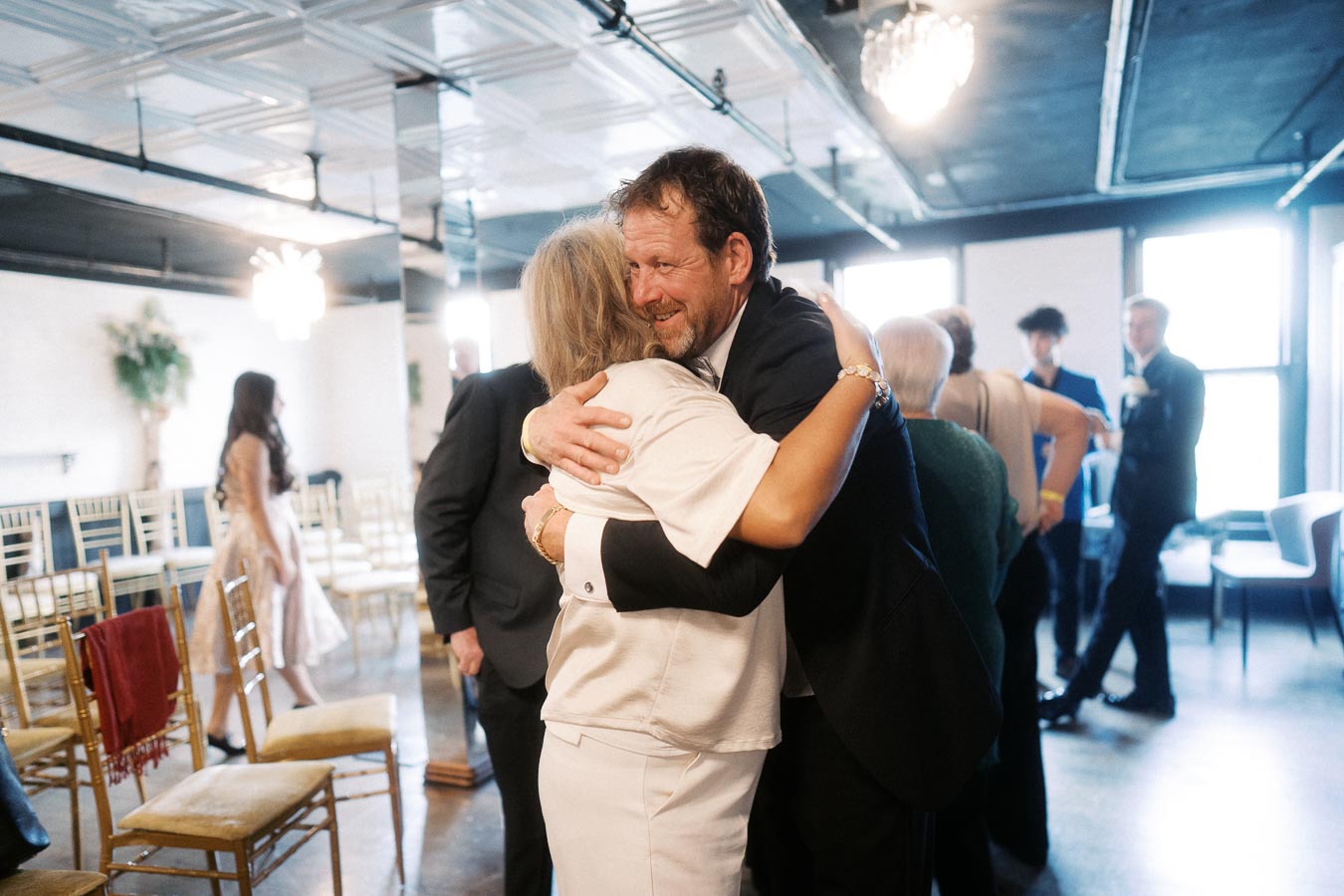 A heartfelt embrace between two people at a lively indoor gathering, surrounded by attendees in formal attire, under elegant lighting.