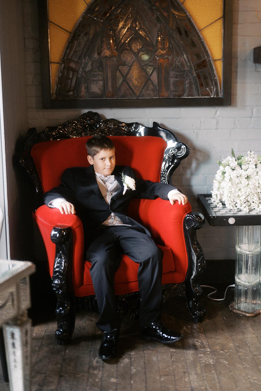 Young boy in a formal suit sitting on an elegant red armchair with ornate black woodwork, in a vintage-themed room with stained glass decor and a floral arrangement on a side table.