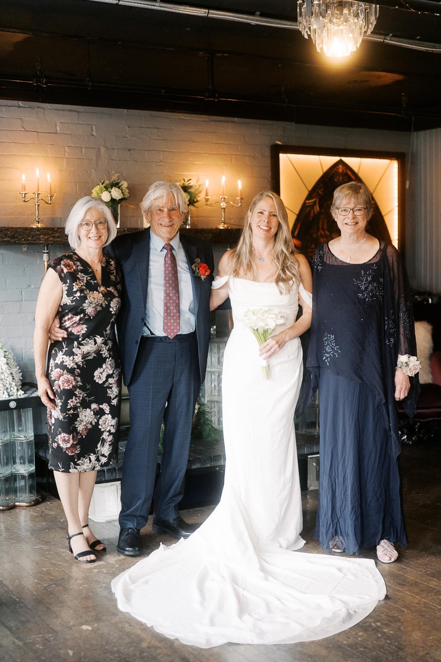 A bride in a white dress poses with three elegantly dressed guests in a warmly lit wedding venue, surrounded by floral arrangements and candles.