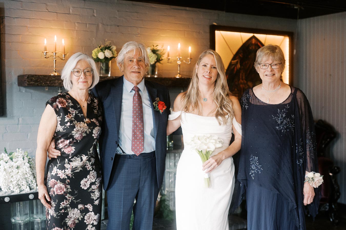 A group of four people, including two women in elegant dresses and one holding a bouquet, stand in a warmly lit room with floral decorations and candles on a shelf.