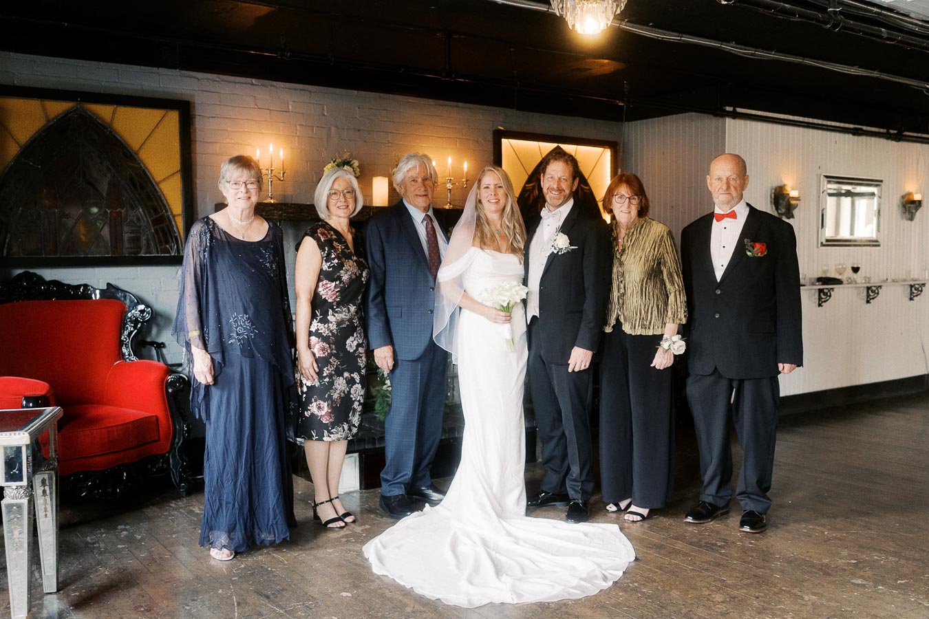 Family wedding portrait with bride and groom in formal attire, surrounded by elegantly dressed family members, posing indoors with decorative background elements and soft lighting.