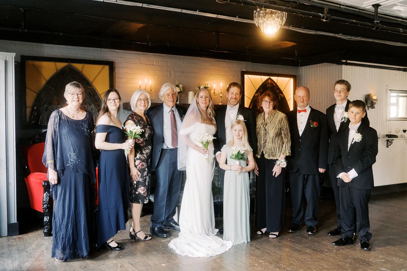 A group of people dressed in formal attire at a wedding ceremony, featuring a bride in a white gown with a veil, a groom in a suit, flower girls, and family members. They are standing indoors with elegant decor, including chandeliers and candelabras, creating a warm and celebratory atmosphere.