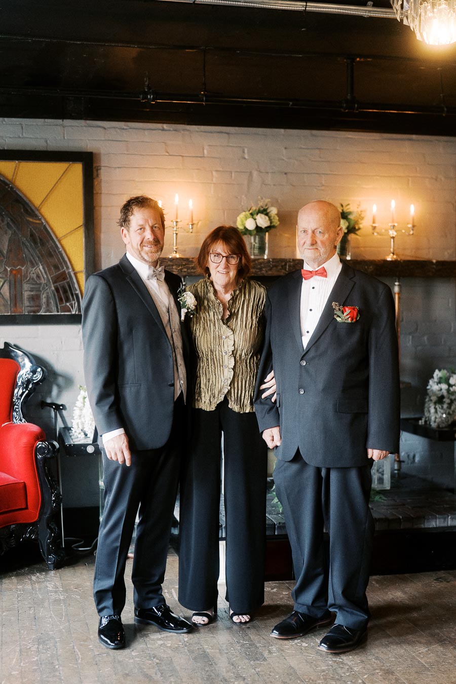 Three elegantly dressed individuals posing indoors with a stylish, sophisticated backdrop of candles and art; two men in suits and a woman in chic evening attire create a formal and warm atmosphere.