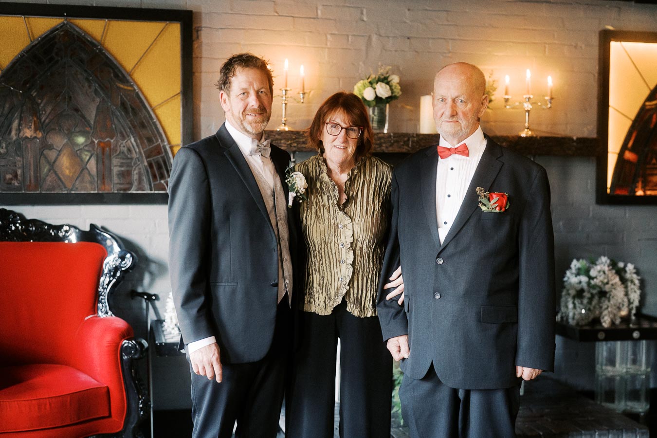 A group of three formally dressed individuals standing together in a warmly lit room with elegant decor, including a red upholstered chair and framed wall art in the background.