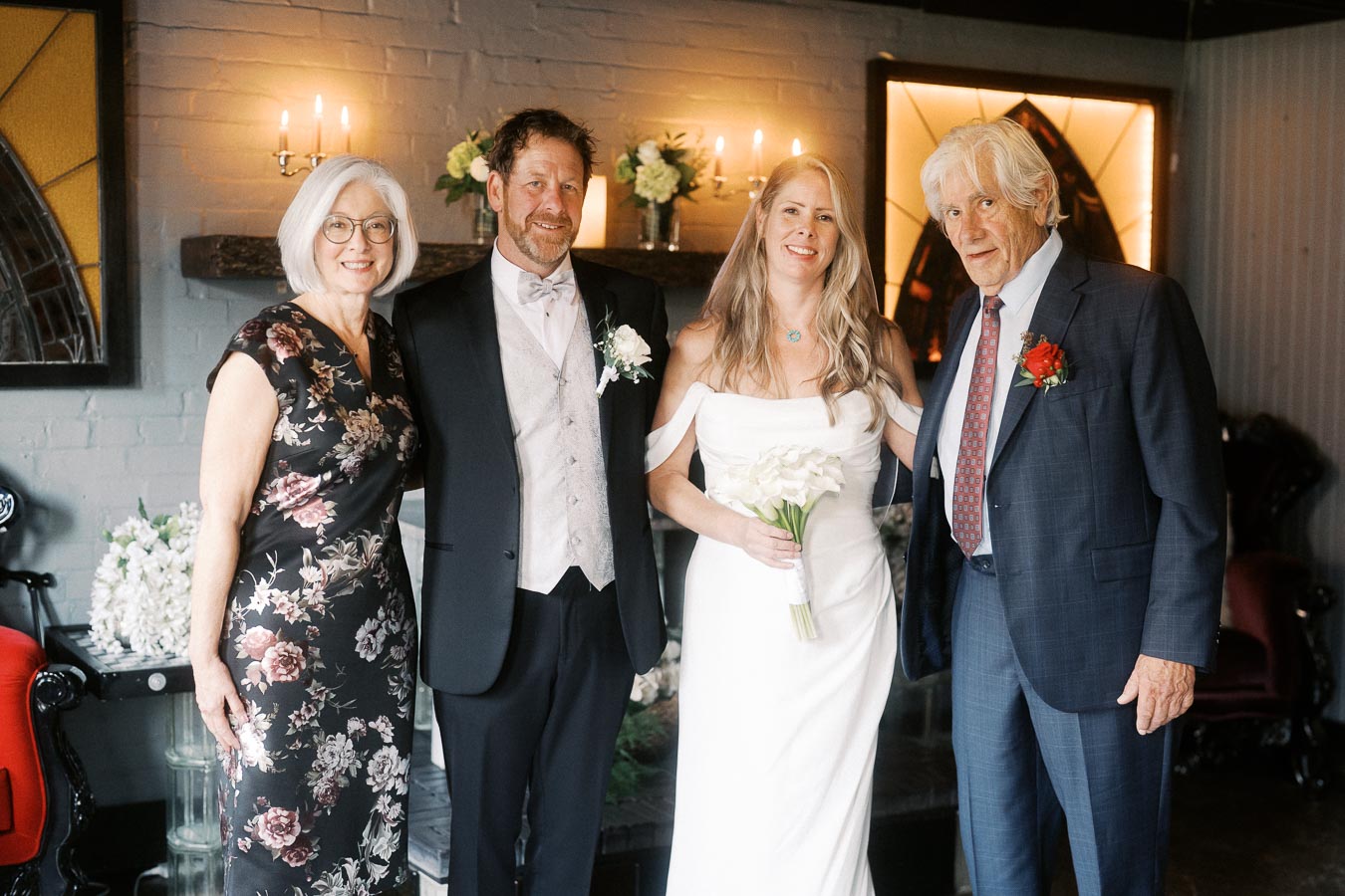 Four people dressed in formal attire posing together indoors, with a bride holding a bouquet. The setting features elegant lighting and floral arrangements, suggesting a wedding or special event.