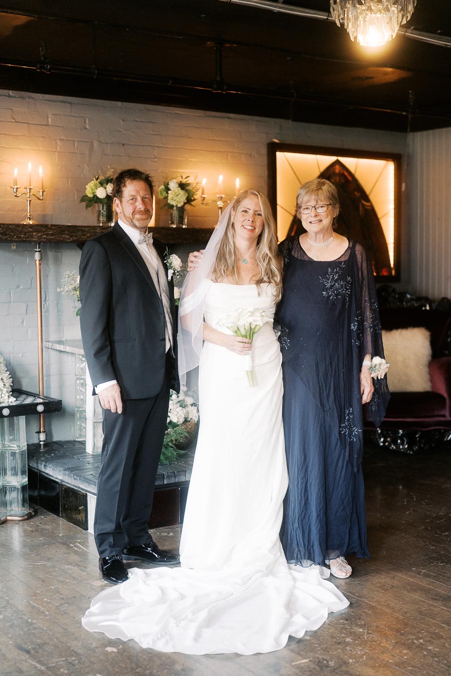Bride in a white wedding dress poses with two guests in formal attire in an elegantly decorated venue with soft lighting and floral arrangements.