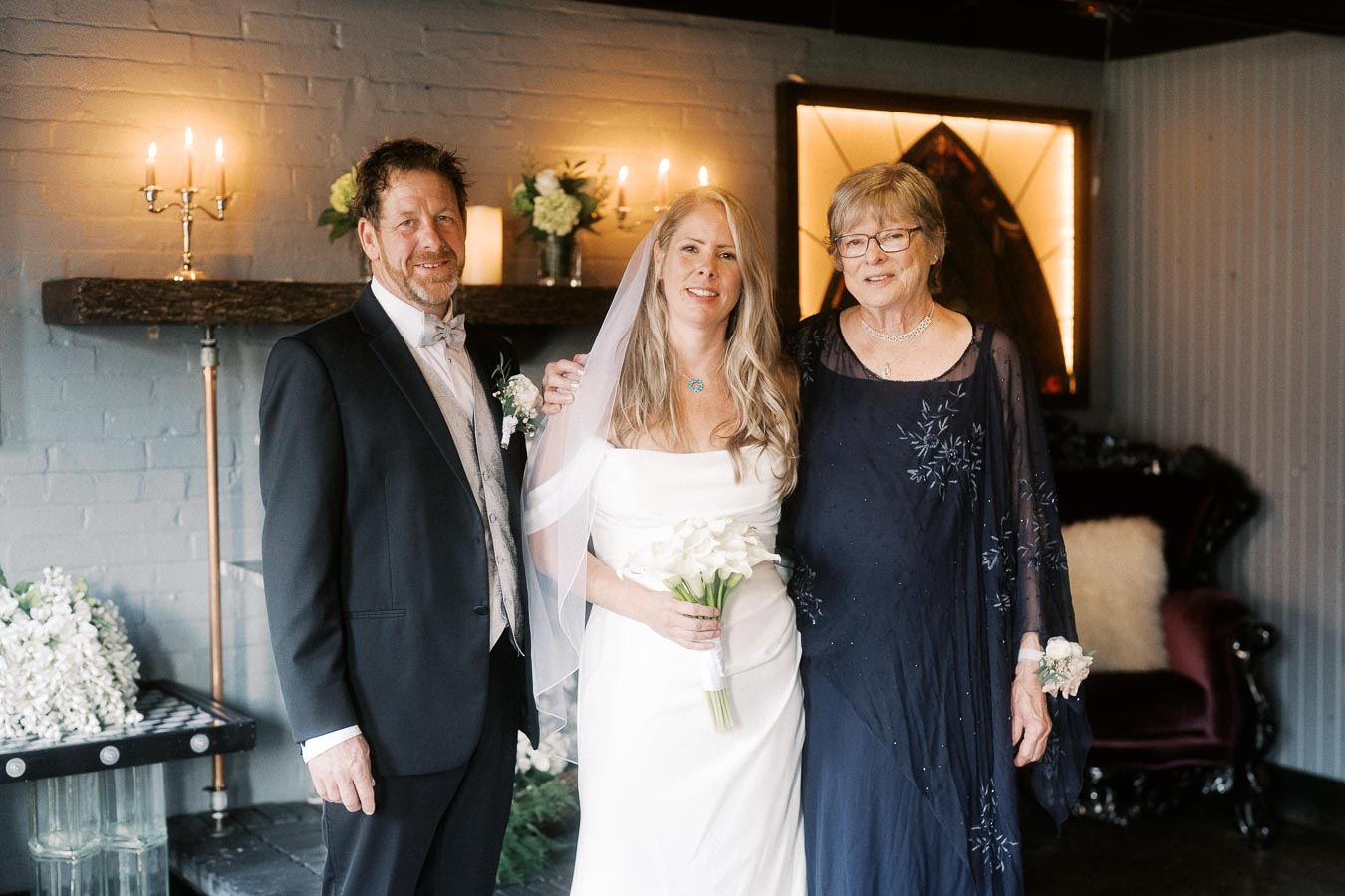 A bride in a white gown holding a bouquet stands between two older adults, all smiling in a warmly lit room with candles and floral arrangements, suggesting a wedding or family celebration atmosphere.
