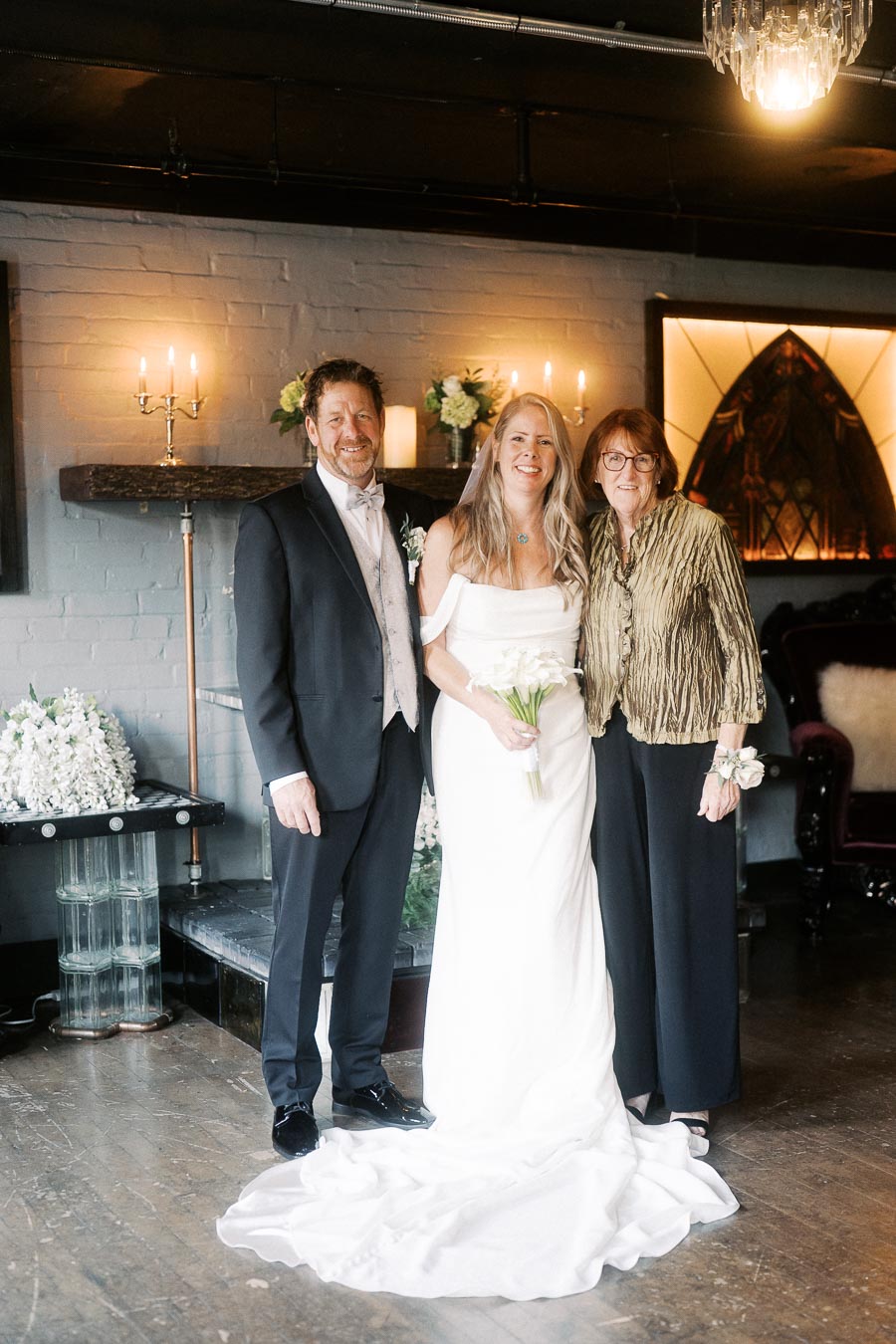 Bride and groom posing with a guest at an indoor wedding ceremony, elegantly decorated with candles and floral arrangements.