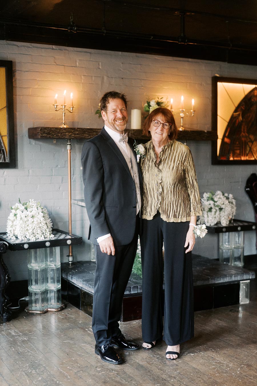 A happy couple elegantly dressed, posing indoors against a stylish backdrop with candles and flower arrangements.