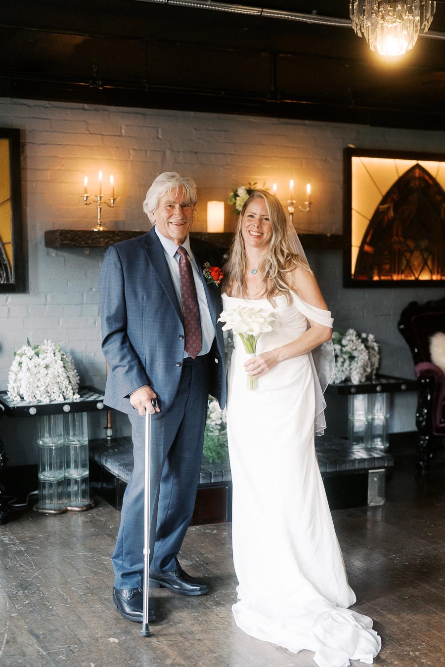 Elderly man in a blue suit and cane standing beside smiling woman in a white wedding dress holding flowers in a warmly lit room with candles and floral decorations.