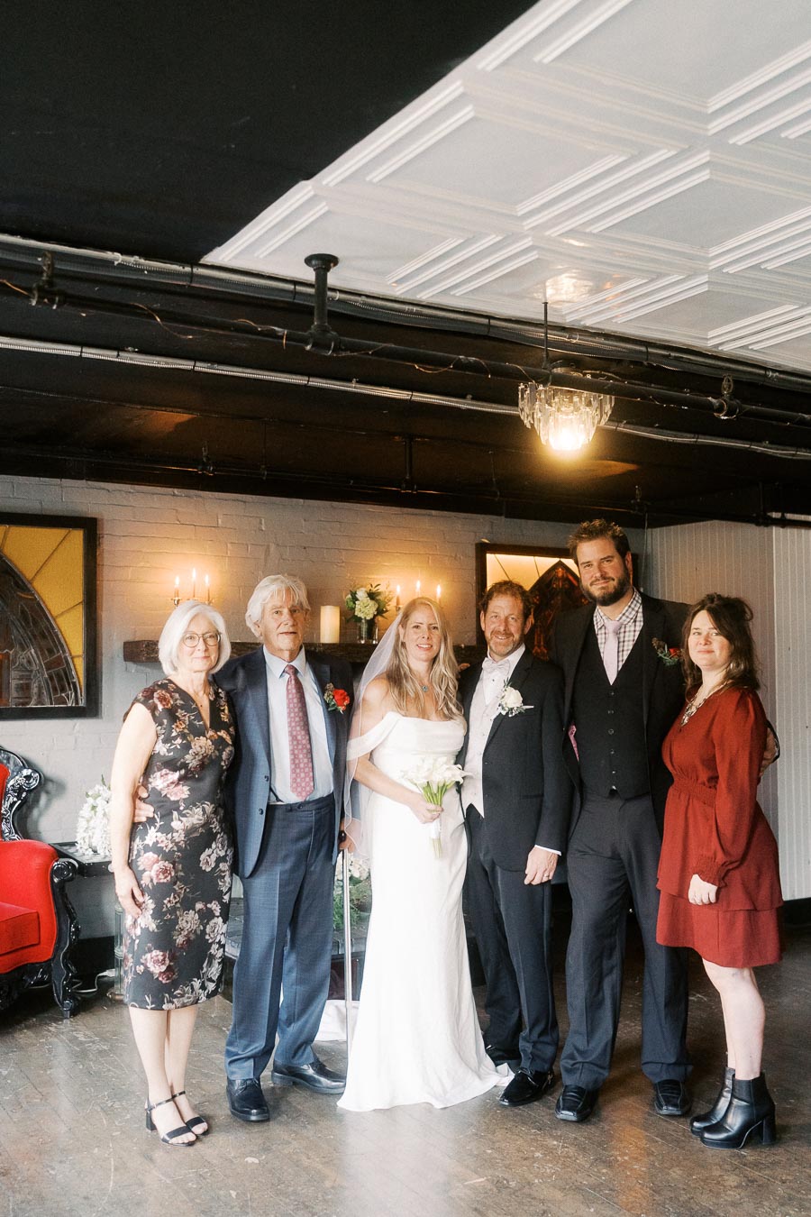 Wedding party group photo in an elegant indoor setting, featuring the bride in a white gown, groom in a suit, and family members in formal attire, standing together with smiles and a warm ambiance.