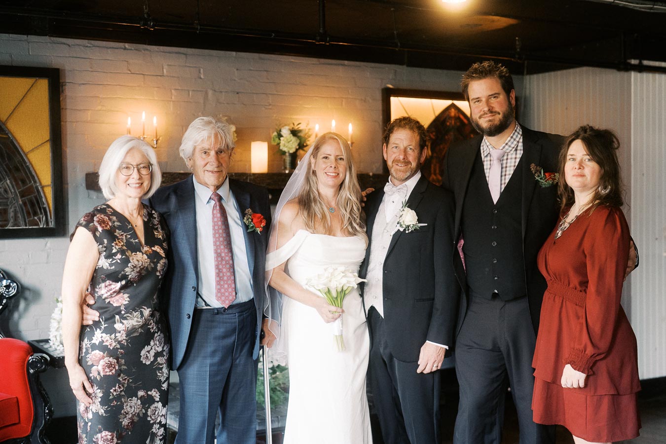 A group of people posing for a wedding photo in a warmly lit room, featuring the bride in a white gown holding a bouquet, surrounded by well-dressed guests, with candles and floral arrangements in the background.
