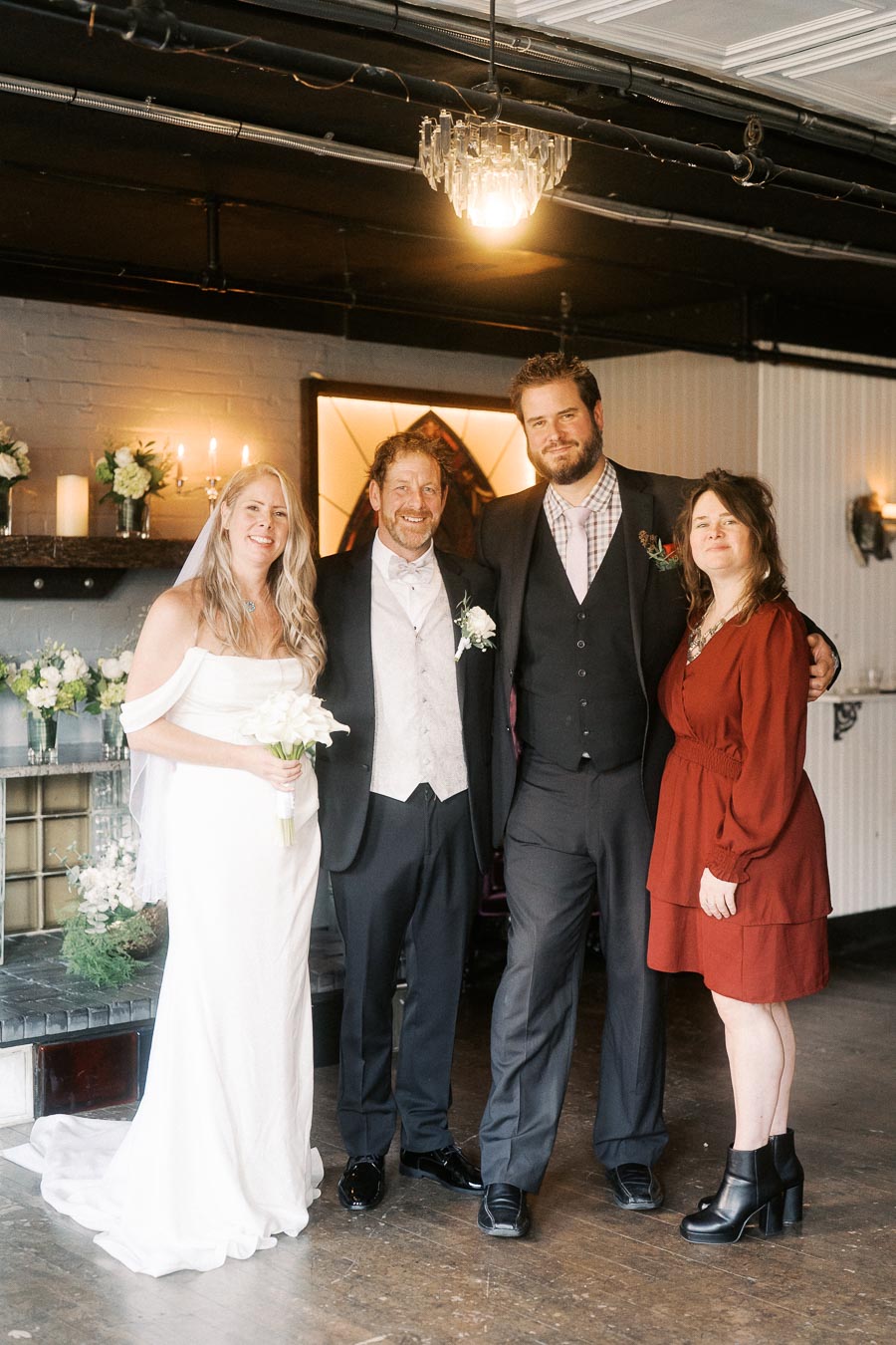 Wedding group photo with bride in white gown holding a bouquet, standing beside groom in formal suit, next to two guests in stylish attire, posing indoors with elegant decor and floral arrangements.