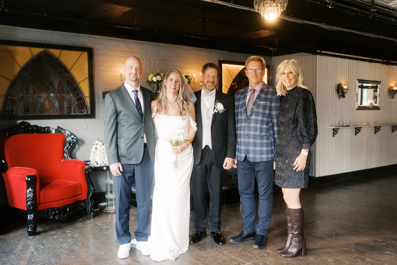 A bride in a white dress holds a bouquet, standing with four guests dressed in formal attire inside a decorated room with a red chair and vintage walls.