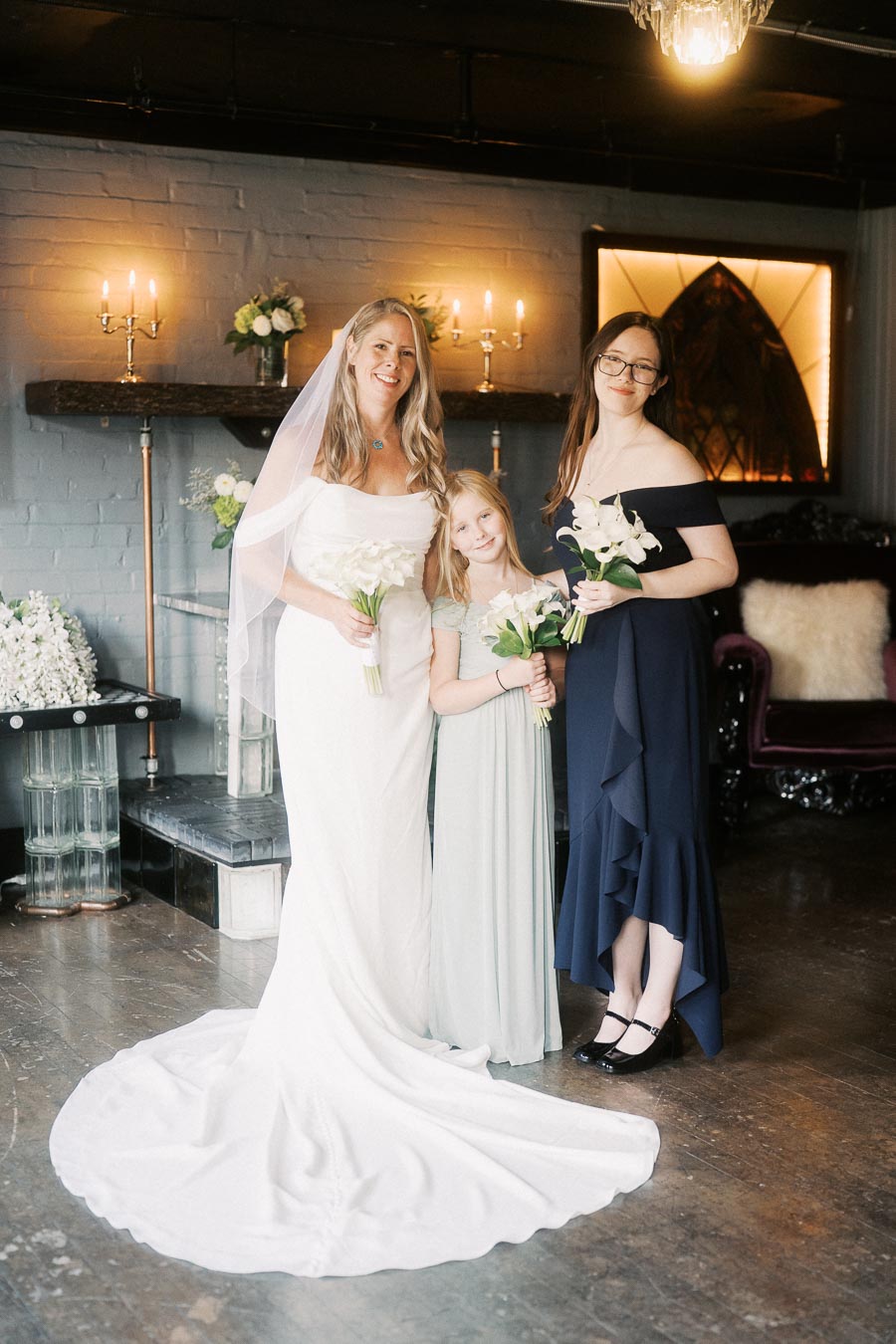 Bride and bridesmaids smiling in a cozy, elegant room with soft lighting, holding white flower bouquets.