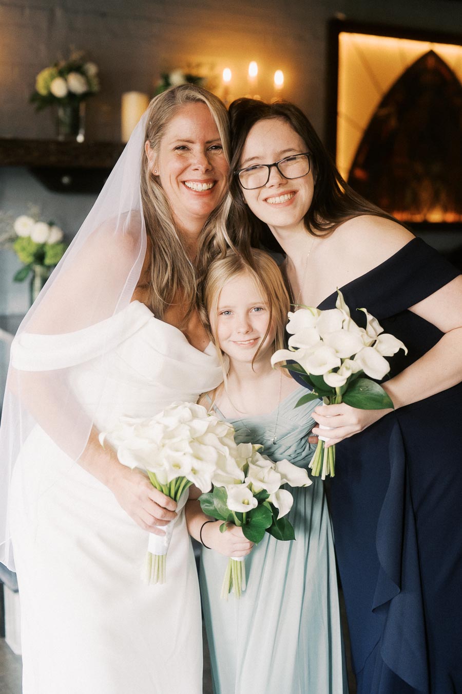 A joyful wedding scene with a bride in a white gown and veil, accompanied by two women, all smiling and holding bouquets of white flowers, set against a softly lit background.