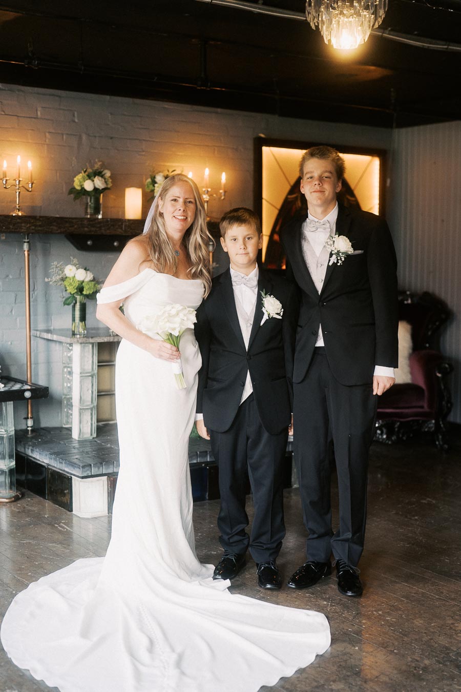 Bride in an elegant white wedding dress standing with two young boys in tuxedos in a warmly lit room, featuring candles and floral decorations.
