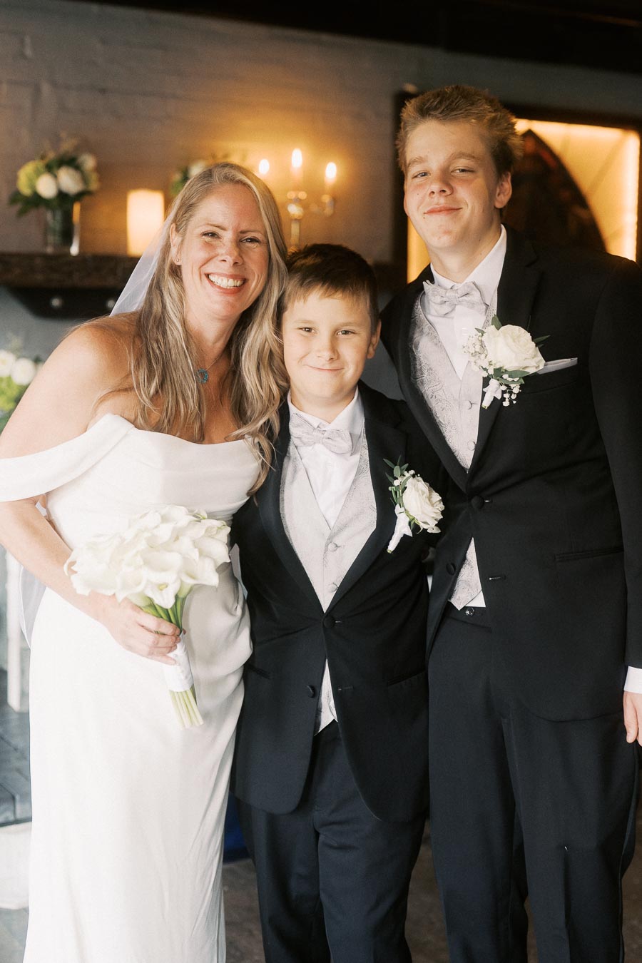 Bride smiling with two young boys in tuxedos at a wedding ceremony. The bride holds a bouquet of white flowers, while everyone is dressed in formal attire, featuring elegant suits and floral boutonnieres. The background has soft lighting and decor, creating a warm and celebratory atmosphere.