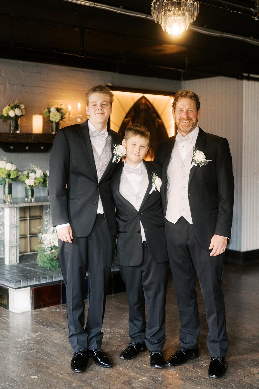 A joyful family moment captured at a wedding, featuring three men dressed in classic black tuxedos with white shirts and matching boutonnieres, standing together in a warmly lit venue adorned with floral arrangements.
