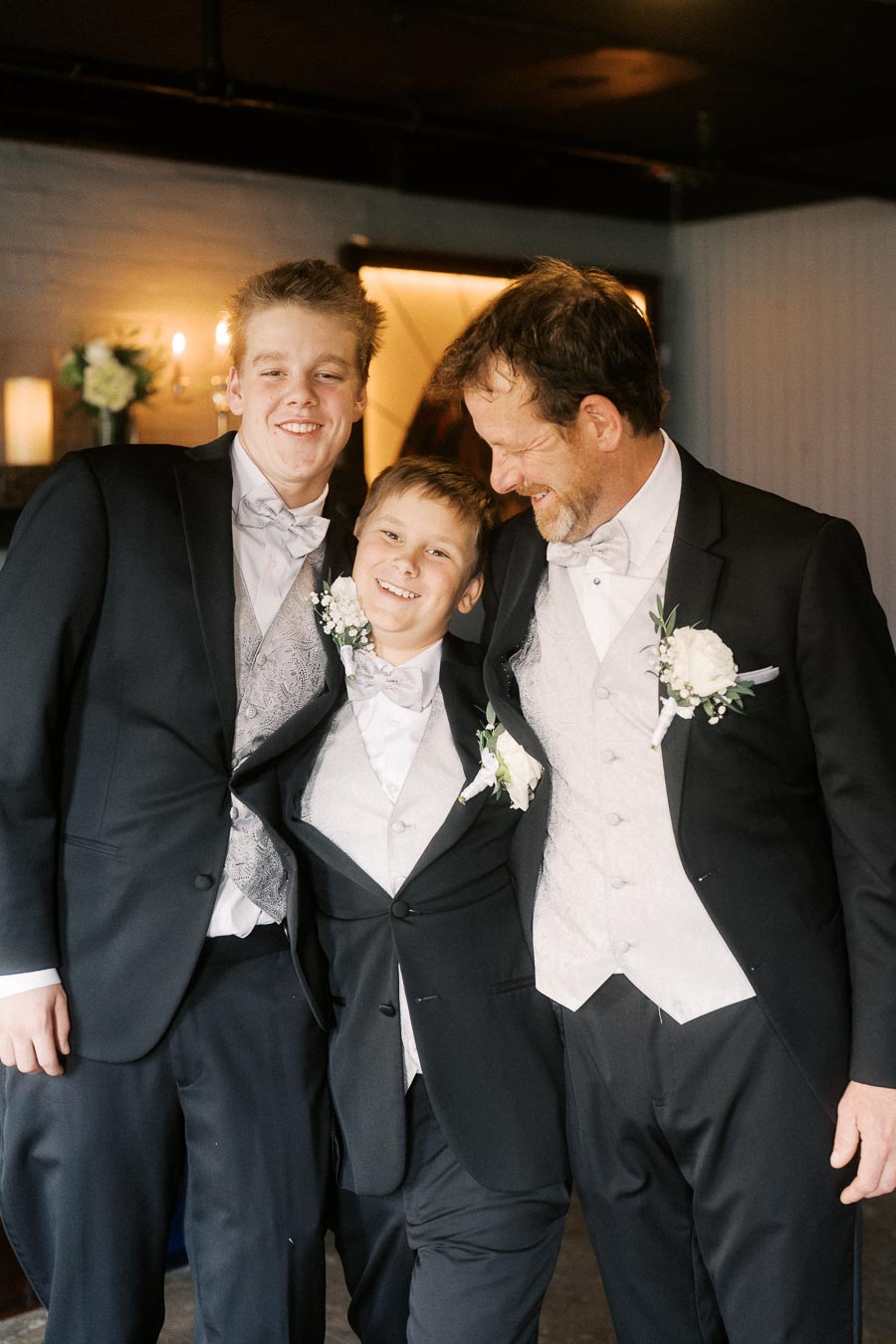 Father and sons in formal attire with boutonnieres, smiling and embracing at a wedding ceremony indoors.