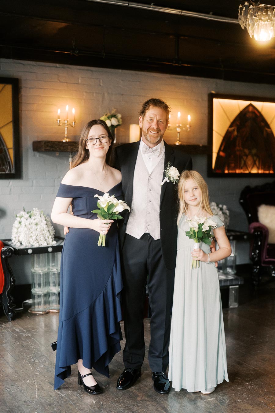A smiling man in formal attire stands with two young girls dressed in elegant dresses holding white flower bouquets in a warmly lit room with decorative candles.